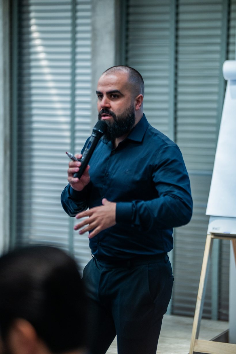Rudnei Mazepa de camisa marinho falando ao microfone durante o evento DOMINA.IA